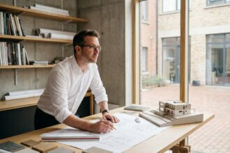 Architecte masculin étudiant des plans de construction détaillés sur une grande table de travail en bois dans un studio professionnel